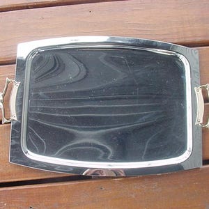 Puede incluir: Una bandeja de servir plateada con dos asas ornamentadas. La bandeja tiene una forma ligeramente curvada y una superficie reflectante.