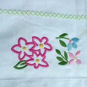 May include: A white fabric textile featuring a horizontal green cross-stitch border at the top. The fabric is decorated with an embroidered cluster of three pink flowers with yellow centers and green leaves, alongside a smaller blue and pink floral motif.