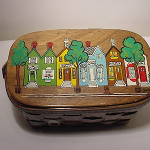 May include: A wooden basket with a painted scene of a row of colorful houses with green trees and a street. The basket has a woven bottom and a hinged lid.