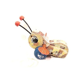 May include: A charming, hand-painted wooden bee ornament with a blue and orange accessory. The bee features a yellow body with brown and white details, black antennae with red tips, and a smiling face. Decorative item.