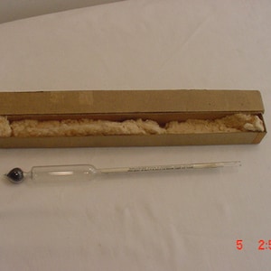Op de afbeelding: Een glazen hydrometer met een zwarte gewichtsbol aan de onderkant, in een kartonnen doos met verpakkingsmateriaal. De hydrometer wordt gebruikt om de soortelijke massa van vloeistoffen te meten.