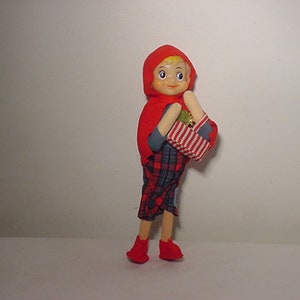 May include: A vintage doll dressed as Little Red Riding Hood, with a red hooded cape, plaid skirt, and red shoes. The doll carries a basket with a red and white striped pattern. The doll has blonde hair and a smiling expression.