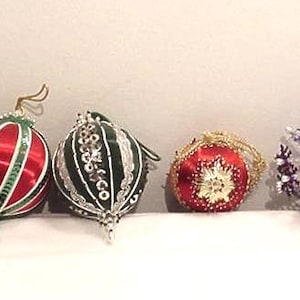 Puede incluir: Cuatro adornos decorativos que incluyen una bola de satén roja y verde con lentejuelas, una bola verde oscuro con adornos plateados, una pequeña bola de satén roja con detalles dorados y un adorno esférico hecho de formas florales de cuentas moradas y blancas.