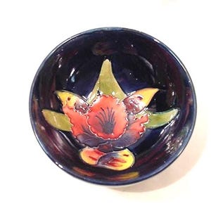 May include: A small, round ceramic bowl with a deep blue interior and a floral design. The flower features red, yellow, and green accents. The bowl's rim is also blue, creating a cohesive and decorative piece.