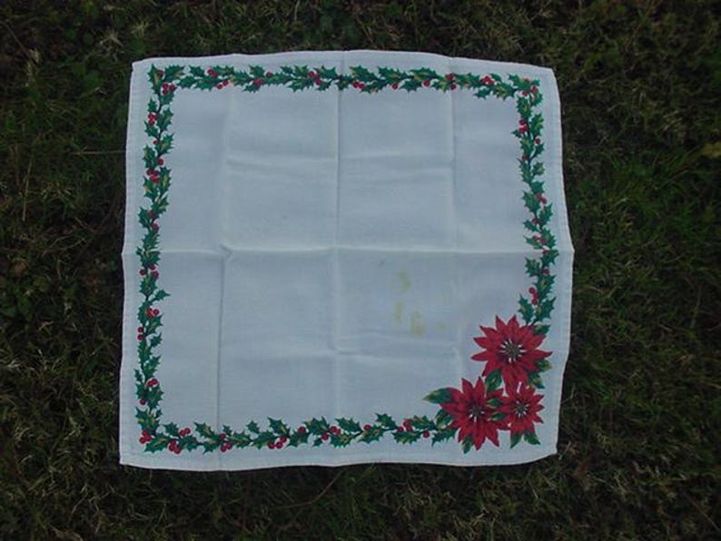 May include: White linen napkin with a green holly and red berry border. The border also features red poinsettia flowers.
