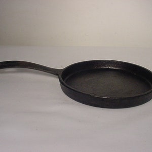 May include: A black cast iron frying pan with a long, curved handle. The pan is round with a slightly raised edge. The skillet is designed for cooking and has a dark, textured surface.