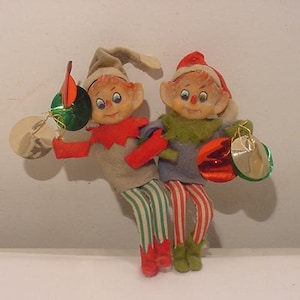 以下が含まれることがあります： 2つのヴィンテージクリスマスエルフオーナメント。1つのエルフは、緑と白のストライプのパンツが付いた赤とタンの衣装を着ています。もう1つのエルフは、赤い帽子と赤と白のストライプのパンツが付いた灰色の衣装を着ています。両方のエルフは装飾的なオーナメントを持っています。