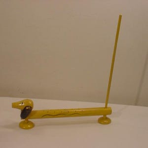 May include: A yellow wooden dachshund incense holder with a black painted nose and eyes. The holder has a long, thin stick extending from the top for incense.