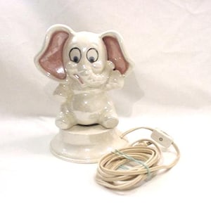 May include: A vintage ceramic elephant lamp in off-white with pink ear accents. The elephant sits on a round base, with a cord and switch. The lamp features large eyes and a raised trunk. A charming decorative item.