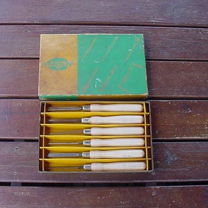 May include: A set of six wood carving chisels with wooden handles, neatly arranged in a yellow and brown box. The box has a green and orange lid with the brand name visible. The tools are displayed on a wooden surface.