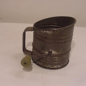 May include: A vintage metal flour sifter with a green plastic handle. The sifter has a worn, rustic look and is made of metal with a white finish.