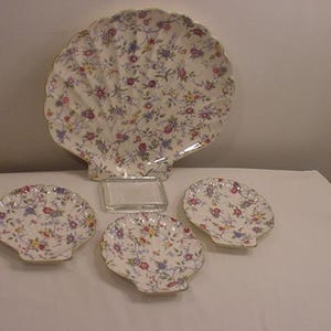 May include: Set of four white ceramic plates shaped like seashells with a floral pattern in shades of pink, blue, and yellow. The plates are arranged on a white surface.