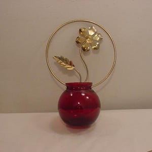 May include: A gold metal flower with a red glass vase. The flower is attached to a gold metal ring.