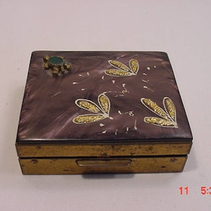 Peut inclure: Un petit compact carré, violet et or, avec un bijou vert sur le dessus. Le compact a un motif floral avec des paillettes dorées.