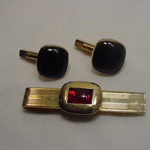 Puede incluir: Un juego de gemelos y un clip para corbata de color dorado. Los gemelos tienen piedras cuadradas negras y el clip para corbata tiene una piedra rectangular roja.