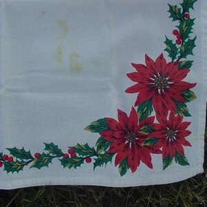May include: White tablecloth with a red and green poinsettia and holly border. The tablecloth has a corner design with three poinsettia flowers and holly leaves.