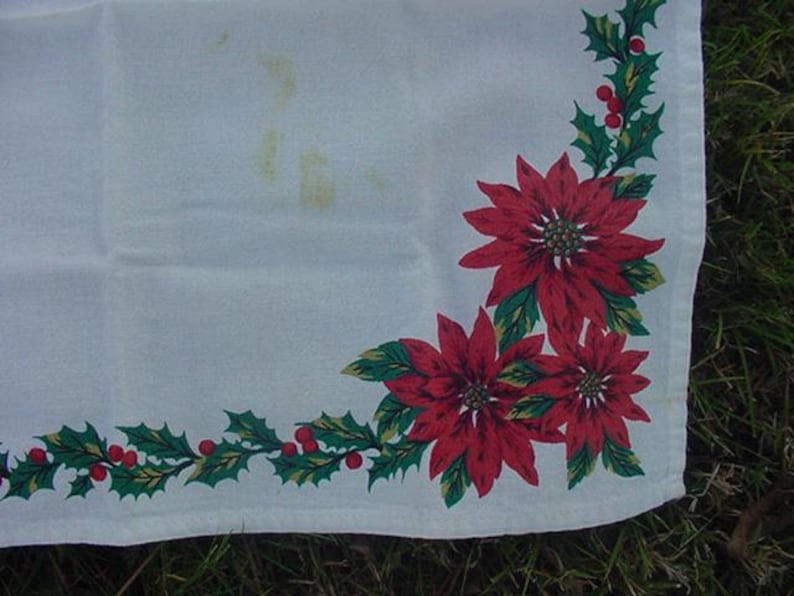 May include: White tablecloth with a red and green poinsettia and holly border. The tablecloth has a corner design with three poinsettia flowers and holly leaves.