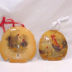 May include: Two decorative ornaments with rooster and hen illustrations. One ornament is a sand dollar shape with a red ribbon, the other is oval with a white ribbon. The ornaments have a glossy finish.