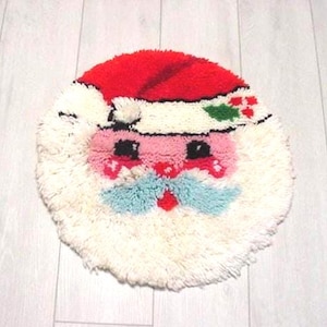 May include: A round, handmade Christmas rug featuring Santa Claus's face. The rug has a red hat, white beard, pink cheeks, and a blue mustache. The design includes black eyes and a holly berry accent. The rug is made of fluffy, textured yarn.