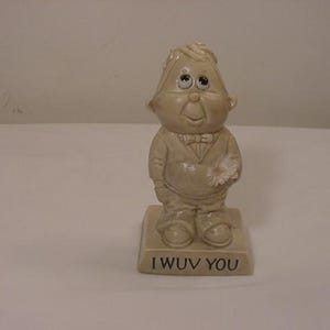 May include: A white ceramic figurine of a person with large eyes and a flower in their hand. The figurine is standing on a base that says "I WUV YOU".