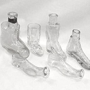 May include: A collection of clear glass boot-shaped bottles and a small glass boot-shaped shot glass. The bottles vary in size and design, with some featuring a stopper. The objects are arranged on a white surface.