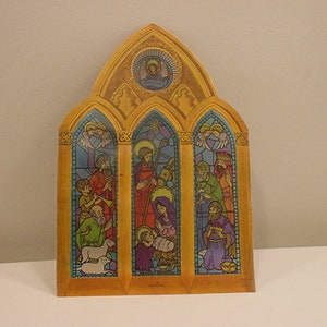 May include: A vintage Christmas card depicting a stained glass window with a nativity scene. The window is divided into three sections, each with a different scene. The center section shows the baby Jesus in a manger, with Mary and Joseph kneeling beside him. The left section shows the three wise men, and the right section shows angels.