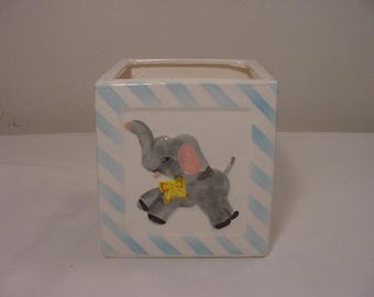 May include: A white ceramic planter with blue and white stripes. The planter features a hand-painted image of a gray elephant with pink ears and a yellow bow.
