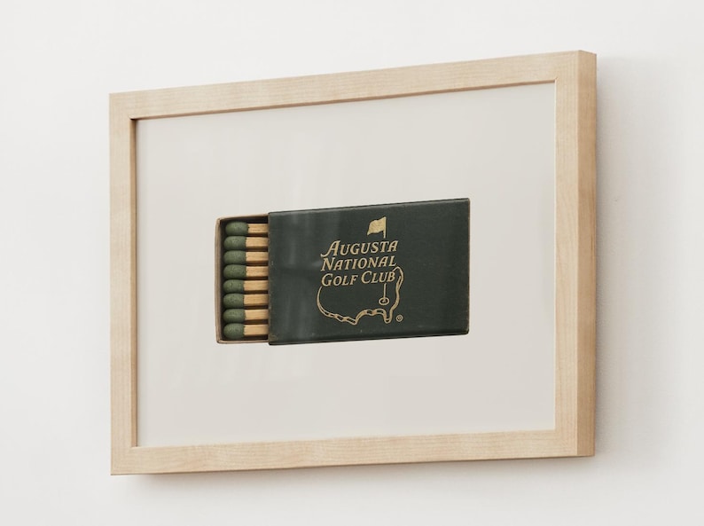 May include: A framed image of a matchbook with a green background and gold lettering. The matchbook says "Augusta National Golf Club" and features a green outline of the golf course. The matchbook is in a wooden frame.