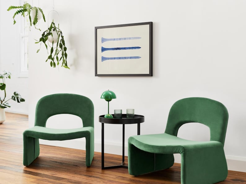 Puede incluir: Dos sillas de terciopelo verde con una mesa auxiliar de metal negro entre ellas. La mesa tiene una l&aacute;mpara verde con una pantalla blanca y dos vasos de vidrio transparente. Una impresi&oacute;n enmarcada con tres l&iacute;neas azules sobre un fondo blanco cuelga de la pared detr&aacute;s de la mesa.