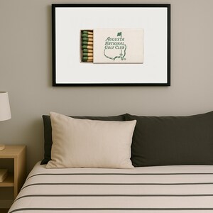 May include: A framed print of a matchbook with the text "Augusta National Golf Club" and a green outline of a golf course. The matchbook has green and white stripes on the side.