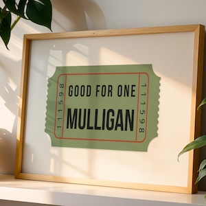May include: A framed print featuring a green ticket design with the words "GOOD FOR ONE MULLIGAN" in bold black letters. The ticket has a red border and a white background, set in a light wood frame.