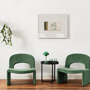 May include: Two green velvet chairs with a black side table in between. The table has a green lamp with a white shade and two clear glass candle holders. A framed print of a matchbook with the text "Augusta National Golf Club" is on the wall behind the table.