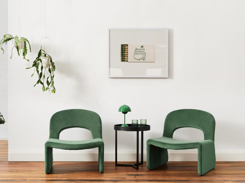 May include: Two green velvet chairs with a black side table in between. The table has a green lamp with a white shade and two clear glass candle holders. A framed print of a matchbook with the text "Augusta National Golf Club" is on the wall behind the table.