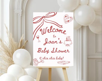 May include: A white sign with red text and illustrations announcing a baby shower for Joan. The sign reads "Welcome to Joan's Baby Shower slice slice baby!" and features illustrations of a bow, pizza, wine glass, a table setting, a scooter, and a book.