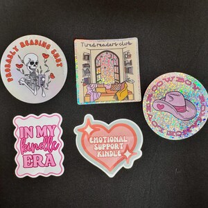 Könnte beinhalten: Eine Sammlung von fünf Aufklebern mit verschiedenen Designs und Texten. Einer zeigt ein Skelett mit dem Text "Probably Reading Shut". Ein anderer sagt "Tired readers club". Weitere Aufkleber sagen "In my Kindle Era", "Emotional Support Kindle" und einen Cowboyhut.