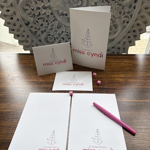 Puede incluir: Un conjunto de papelería blanca que incluye una tarjeta plegada, una tarjeta pequeña y dos hojas de papel, todas con un dibujo lineal negro de un pastel y el texto A note from miss cyndi en rosa y azul, junto a un bolígrafo rosa.