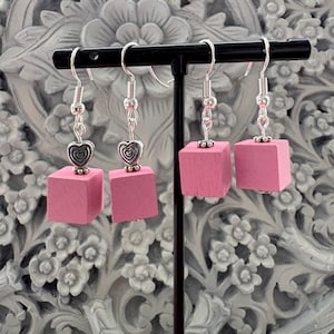 May include: Four pairs of pink cube earrings with silver-colored hooks and heart charms. The earrings are displayed on a black stand against a gray floral background. The earrings have a simple, geometric design.