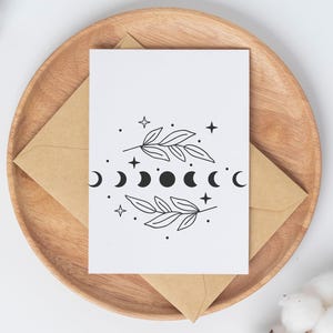 May include: A white card with a black and white illustration of a moon phase cycle with two branches of leaves on either side. The card is sitting on a wooden plate with a brown envelope.