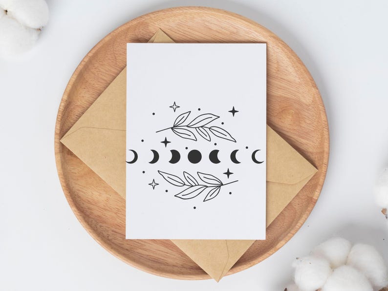 May include: A white card with a black and white illustration of a moon phase cycle with two branches of leaves on either side. The card is sitting on a wooden plate with a brown envelope.