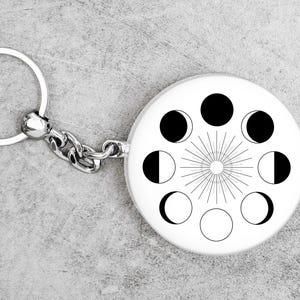 May include: A silver keychain with a round, white charm featuring a black and white moon phase design. The charm has a sunburst design in the center.