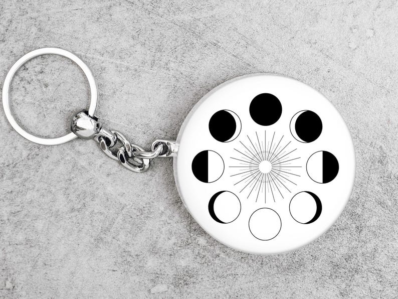 May include: A silver keychain with a round, white charm featuring a black and white moon phase design. The charm has a sunburst design in the center.