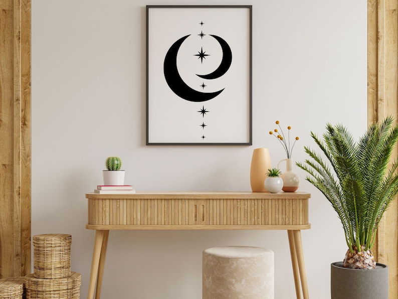 May include: A wooden console table with a light wood finish and a white stool. The table has a black and white print of a crescent moon and stars hanging above it. There are two potted plants on the table, one with a cactus and the other with a palm tree.