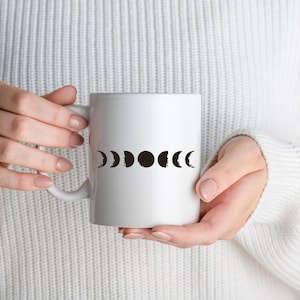 May include: White ceramic mug with a black moon phase design. The mug is being held by a person wearing a white sweater.