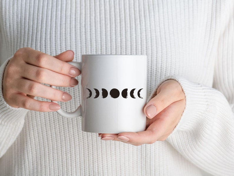 May include: White ceramic mug with a black moon phase design. The mug is being held by a person wearing a white sweater.