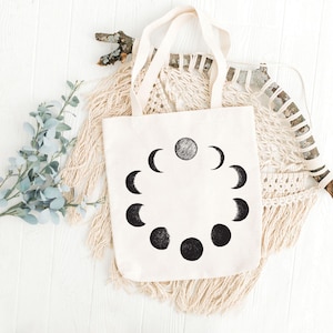 May include: A white canvas tote bag with black moon phases printed on it. The bag has two handles and is lying on a white surface with a macrame wall hanging and a branch behind it.