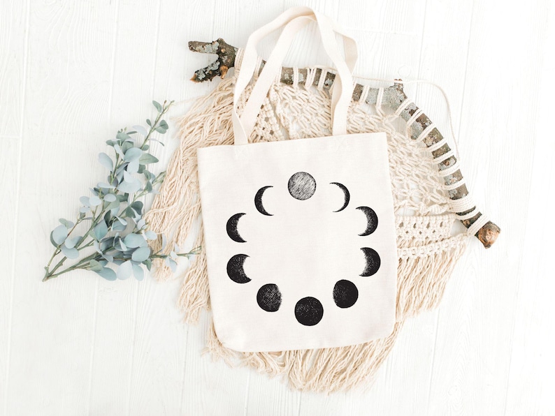 May include: A white canvas tote bag with black moon phases printed on it. The bag has two handles and is lying on a white surface with a macrame wall hanging and a branch behind it.