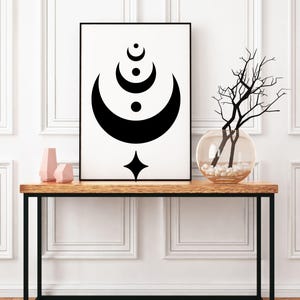 May include: Black and white abstract art print of a crescent moon with a star and circles. The print is framed and displayed on a wooden console table with a vase of branches.