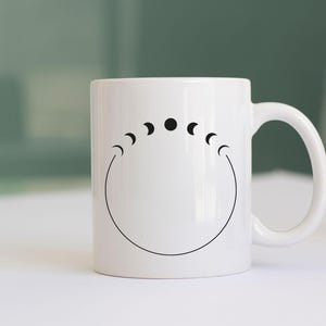 May include: White ceramic mug with a black moon phase design. The design features a circle with five moon phases, including a full moon.