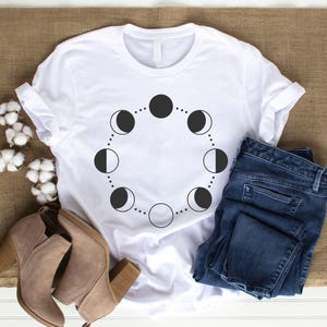 May include: White t-shirt with a black moon phases design. The shirt is laid flat on a brown burlap surface with a pair of brown ankle boots and blue jeans.