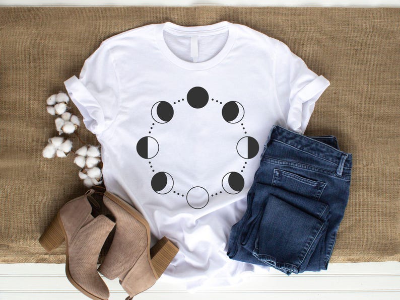 May include: White t-shirt with a black moon phases design. The shirt is laid flat on a brown burlap surface with a pair of brown ankle boots and blue jeans.